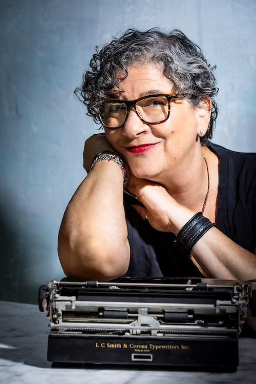 Linda Epstein has light skin tones and pepper-salt curly hair she's wearing heavy tortoise shell eyeglasses and red lipstick, and diamond stud earring. Leaning against her right arm, and on a vintage black typewriter. Wearing beaded bracelets on right wrist and leather cuff on left wrist wearing beaded necklace
